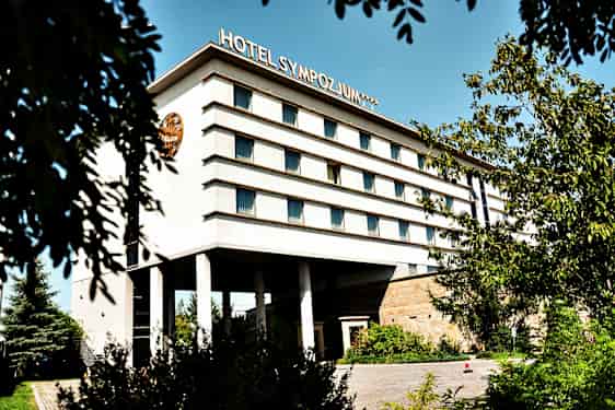 Byggnad med flera våningar, märkt med texten 'Hotel Sympozjum', omgiven av träd och växtlighet. (AI-genererad text)