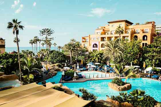 Hotellområde med pooler och grönska framför en byggnad, hav i bakgrunden. (AI-genererad text)