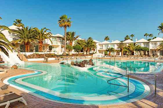 En hotellpool omgiven av palmer, solstolar och vita byggnader. (AI-genererad text)