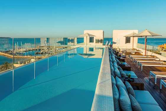 Utsikt över en infinitypool med solstolar och paraplyer, omgiven av glasräcken och en marina i bakgrunden. (AI-genererad text)