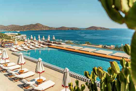 En pool med solstolar och parasoller vid en kustlinje med utsikt över ett blått hav och berg. (AI-genererad text)