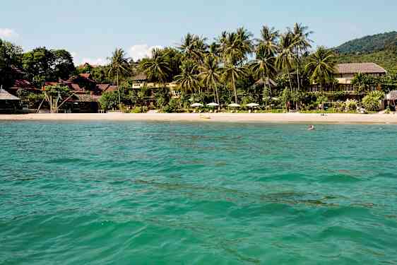 Vy över en sandstrand omgiven av tropiska träd och byggnader, sedd från vattnet. (AI-genererad text)