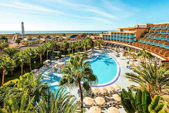 En hotellanläggning med en stor pool, solstolar och parasoller omgiven av palmer, med havet och en fyr i bakgrunden. (AI-genererad text)