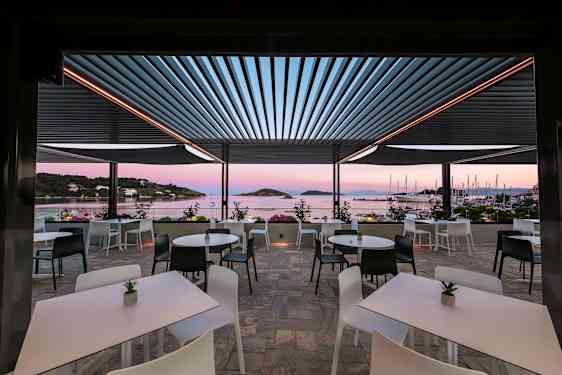 En utomhusterrass med bord och stolar med utsikt mot havet och segelbåtar. (AI-genererad text)