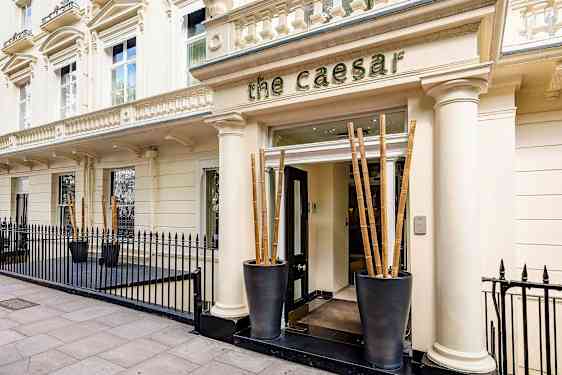 Fasad med ingång till hotell The Caesar, omgiven av svart järnstaket. (AI-genererad text)