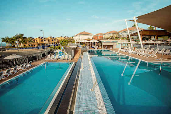 En bild av en solig semesterort med två utomhuspooler omgivna av solstolar, med byggnader i bakgrunden. (AI-genererad text)