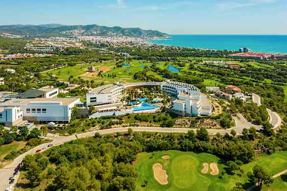 Vy från ovan av ett hotellkomplex omgivet av grönska och en golfbana. Havet och en stad ligger i bakgrunden. (AI-genererad text)