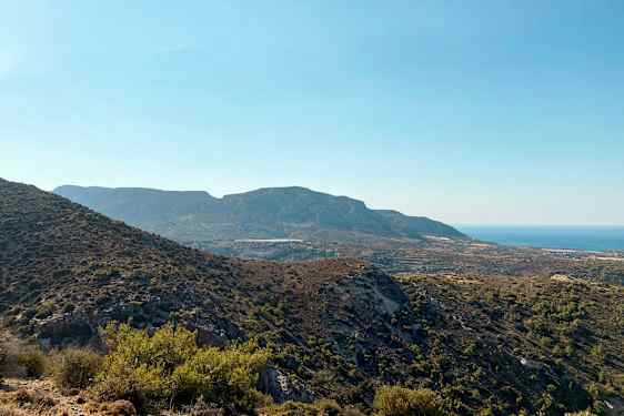 En vy över ett bergigt landskap med varierande växtlighet och havet i bakgrunden under en klarblå himmel. (AI-genererad text)
