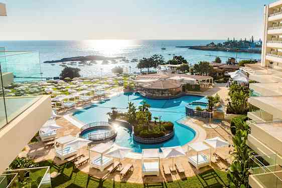 Bilden visar en utomhuspool vid ett hotellkomplex nära havet, omgivet av solstolar, parasoller och grönska. (AI-genererad text)