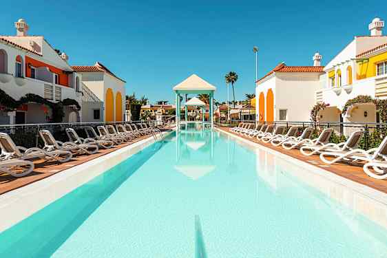 En utomhuspool omgiven av solstolar och färgglada byggnader med en klar himmel. (AI-genererad text)