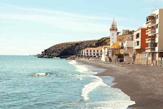 Bild på en kust med svarta sandstränder, en fyr och byggnader vid vattenkanten. (AI-genererad text)