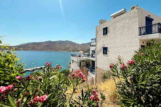 Vit byggnad med blå detaljer vid havet, omgiven av buskar med rosa blommor. (AI-genererad text)