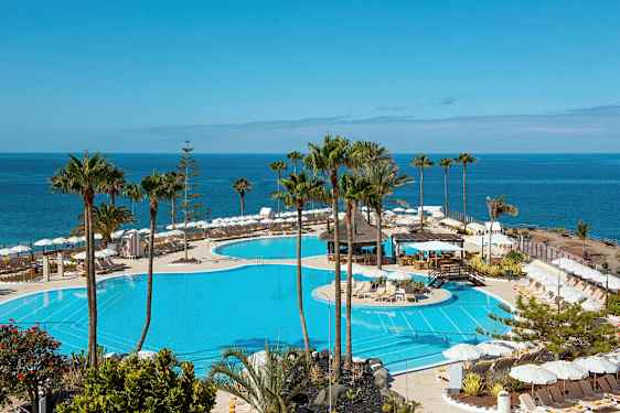 Utsikt över en stor pool med solstolar och parasoller runt omkring, samt havet i bakgrunden. (AI-genererad text)