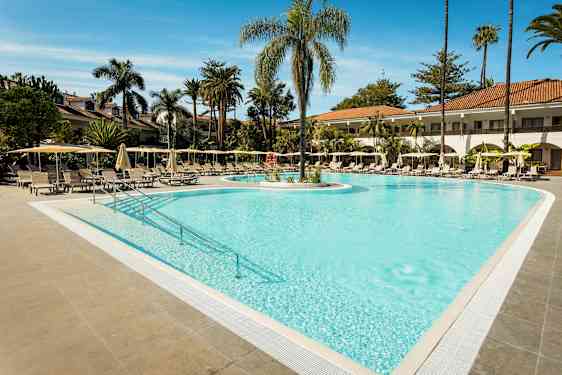 En utomhuspool omgiven av solstolar och parasoller med palmträd i bakgrunden. (AI-genererad text)