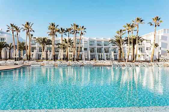 Hotellets poolområde med klarblått vatten, omgivet av solstolar, parasoller och palmträd. I bakgrunden syns en vit byggnad med balkonger. (AI-genererad text)