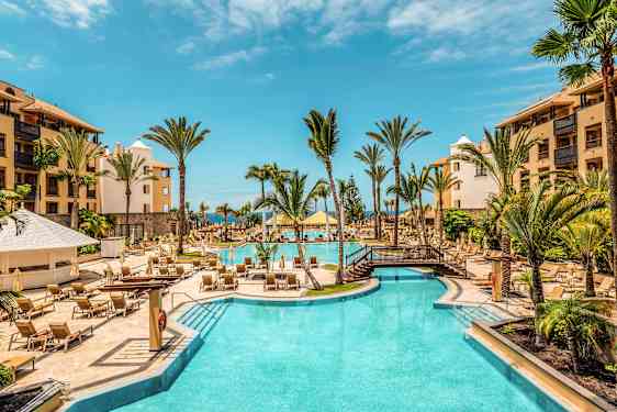 Hotellpool utomhus med palmer och solstolar, omgiven av byggnader. (AI-genererad text)