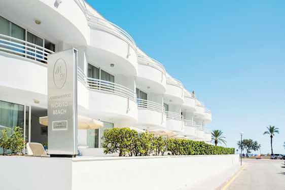 Den vita byggnaden tillhör Morito Beach Apartments, med palmer och hav i bakgrunden. (AI-genererad text)