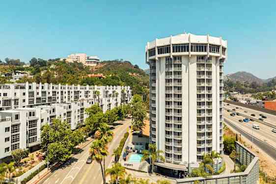 Ett modernt höghus omgiven av vägar, grönska och andra byggnader. (AI-genererad text)