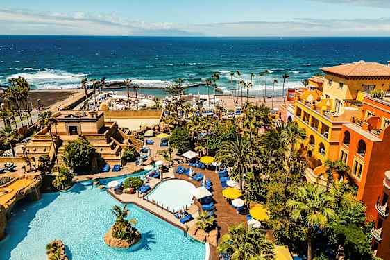 Ett hotellområde med pooler, växtlighet och färgglada byggnader vid havet. (AI-genererad text)