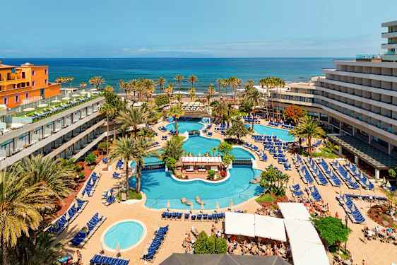 Det är en utsikt över ett hotellområde med flera pooler, omgivna av solstolar och parasoller, samt träd och byggnader. I bakgrunden syns havet. (AI-genererad text)