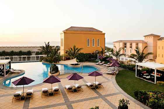 En utomhuspool med solstolar och parasoller ligger omgiven av palmer och en byggnad med gula fasader. (AI-genererad text)