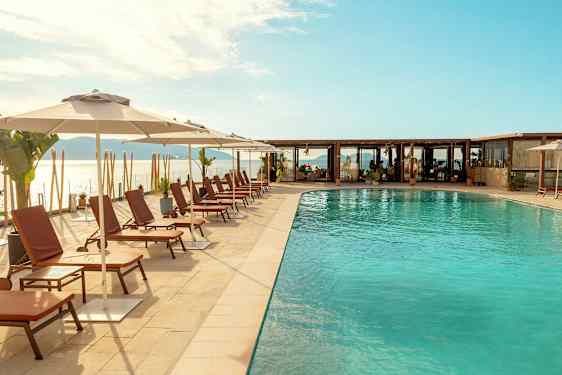 Bilden visar en utomhuspool med solstolar och parasoller vid poolkanten, med hav och molnig himmel i bakgrunden. (AI-genererad text)