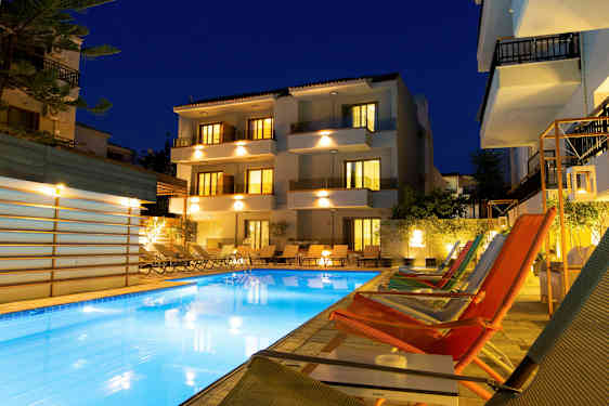 Bilden visar en upplyst hotellbyggnad och en utomhuspool omgiven av solstolar och loungestolar under kvällstid. (AI-genererad text)