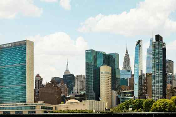 Bilden visar en stadssiluett med många höga byggnader och skyskrapor omgivna av grönska under en klar himmel. (AI-genererad text)