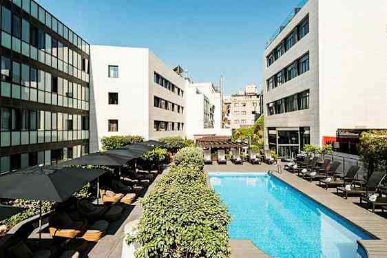 En hotellpool omgiven av liggstolar och parasoller, med moderna byggnader runtom. (AI-genererad text)