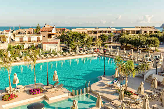 En utomhuspool omgiven av solstolar och parasoller, med byggnader och havet i bakgrunden. (AI-genererad text)