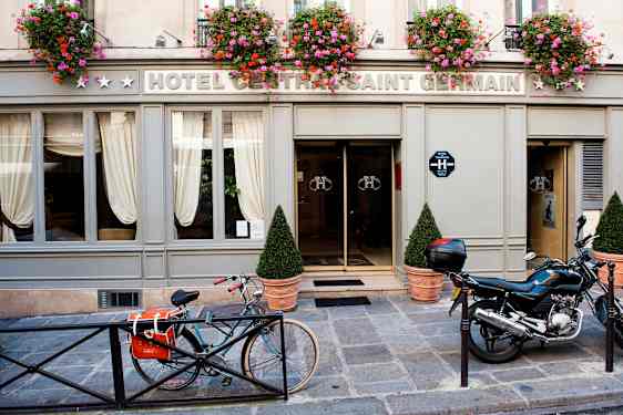 Exteriören av ett hotell med skylten 'Hotel Central Saint Germain' med blomkrukor ovanför ingången samt en cykel och motorcykel parkerad framför. (AI-genererad text)