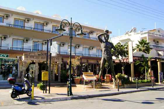 Staty framför byggnad med butiker och restaurang. En scooter står parkerad intill. (AI-genererad text)