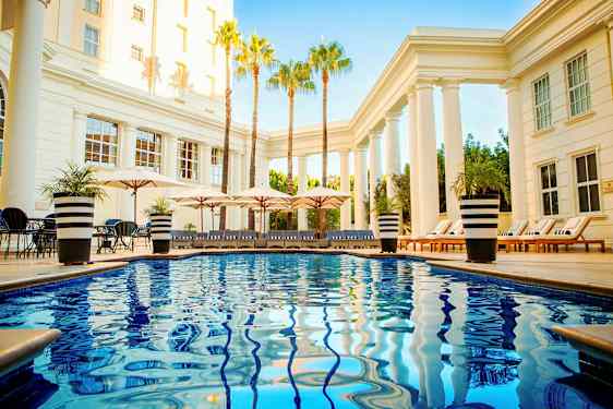 Poolområde med blanka blåa vatten, solstolar och parasoller omgivna av pelare och palmer. (AI-genererad text)