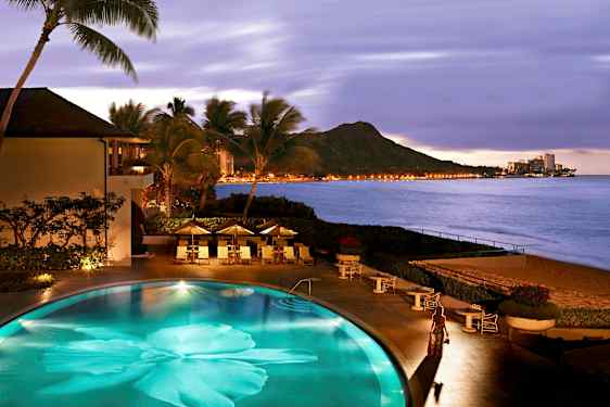 En pool vid stranden omgiven av palmer, med utsikt över berg och ljusen från en stad vid havet. (AI-genererad text)