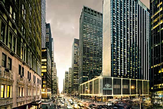 En stadsvy med höga skyskrapor, livlig trafik och byggnader med många fönster. Hilton-hotellet syns längst ner. (AI-genererad text)