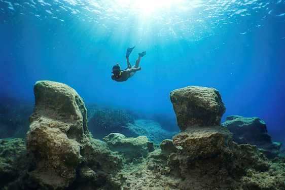 En snorklare simmar under vattnet mellan stora klippor med solljus som filtreras ner från ytan. (AI-genererad text)