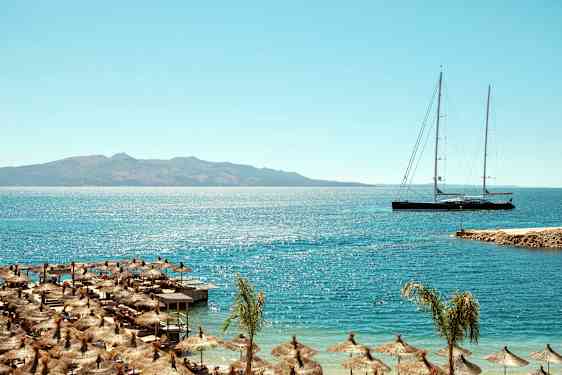 Strand med parasoller och lång segelbåt på havet med berg i bakgrunden. (AI-genererad text)
