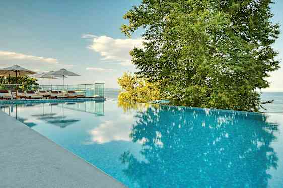 En infinitypool med solstolar och parasoller bredvid, omgiven av träd och med utsikt över havet. (AI-genererad text)