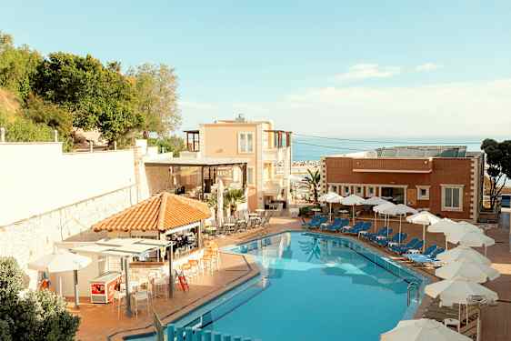 En hotellpool med solstolar och parasoller vid sidan om samt byggnader och grönska i bakgrunden. (AI-genererad text)