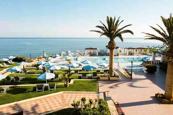 Ett hotellområde med en pool och solstolar under blå parasoller, omgärdad av palmer och med utsikt över havet. (AI-genererad text)