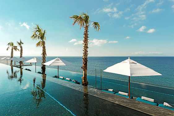 En infinitypool med utsikt mot havet, omgiven av solparasoller och palmer under en klarblå himmel. (AI-genererad text)