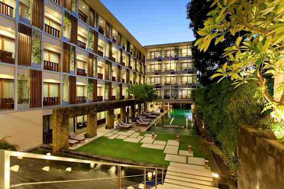 Ett hotellområde under kvällstid med upplysta fönster, en rektangulär pool omgiven av gröna växter och solstolar samt en välskött gräsmatta. (AI-genererad text)