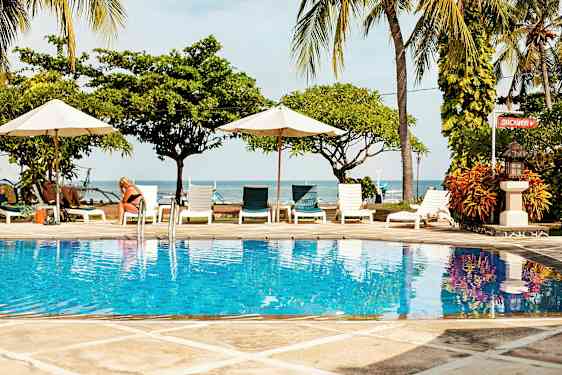 Bild av en pool med klart blått vatten, omgiven av solstolar och parasoller med havet samt palmträd i bakgrunden. (AI-genererad text)