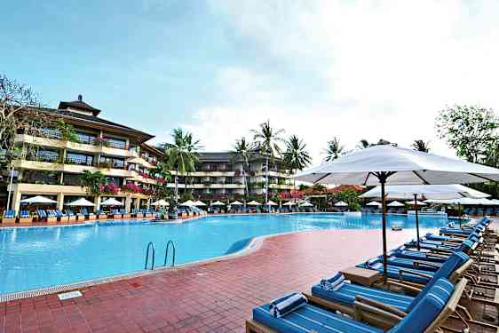 Bild av en utomhuspool omgiven av solstolar och parasoller, med ett fler vånings hotell och palmer i bakgrunden. (AI-genererad text)