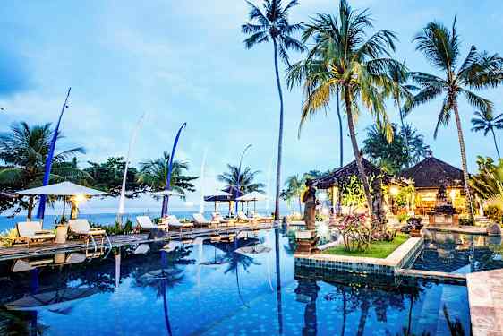 En infinitypool omgiven av solsängar, parasoller, höga palmer och ett öppet paviljongområde vid skymning. (AI-genererad text)