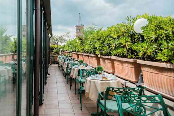 En utomhusterrass med dukade bord och gröna metallstolar, kantad av stora planteringslådor med gröna växter. (AI-genererad text)