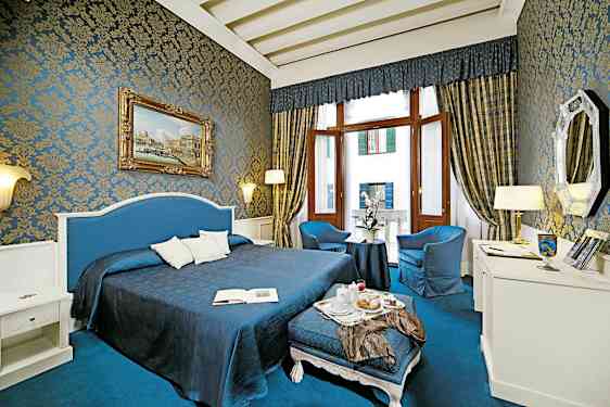 Ett vackert inrett hotellrum med blå och gyllene detaljer, stor säng, sittgrupp och stora fönster. (AI-genererad text)