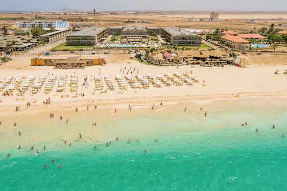 Flygfoto av en strand med turkost vatten, solstolar och parasoller, samt bakom en hotellbyggnad. (AI-genererad text)