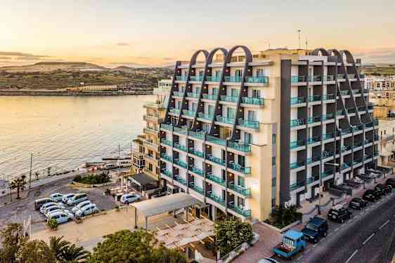 En byggnad vid vattnet med moderna våningsplan och balkonger. I förgrunden syns parkerade bilar och en väg. (AI-genererad text)