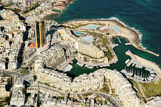 Flygfoto över en kuststad med höga byggnader, småbåtshamn och havet i bakgrunden. (AI-genererad text)